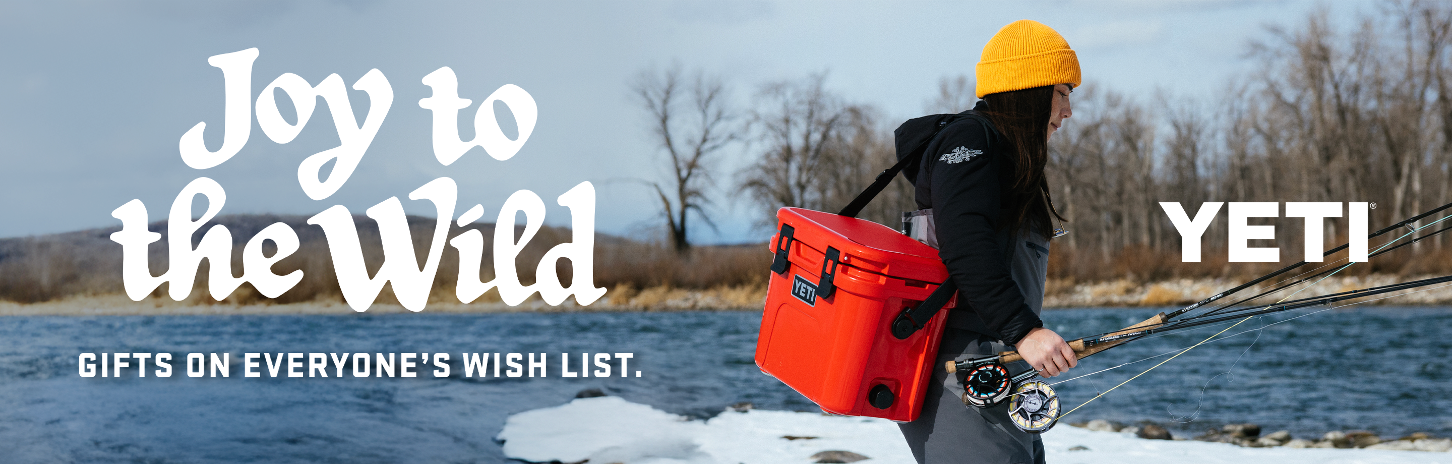 A woman carrying a Yeti cooler on her way to fish an icy river; text reads, "Joy to the Wild, Gifts on Everyone's Wish List — Yeti"