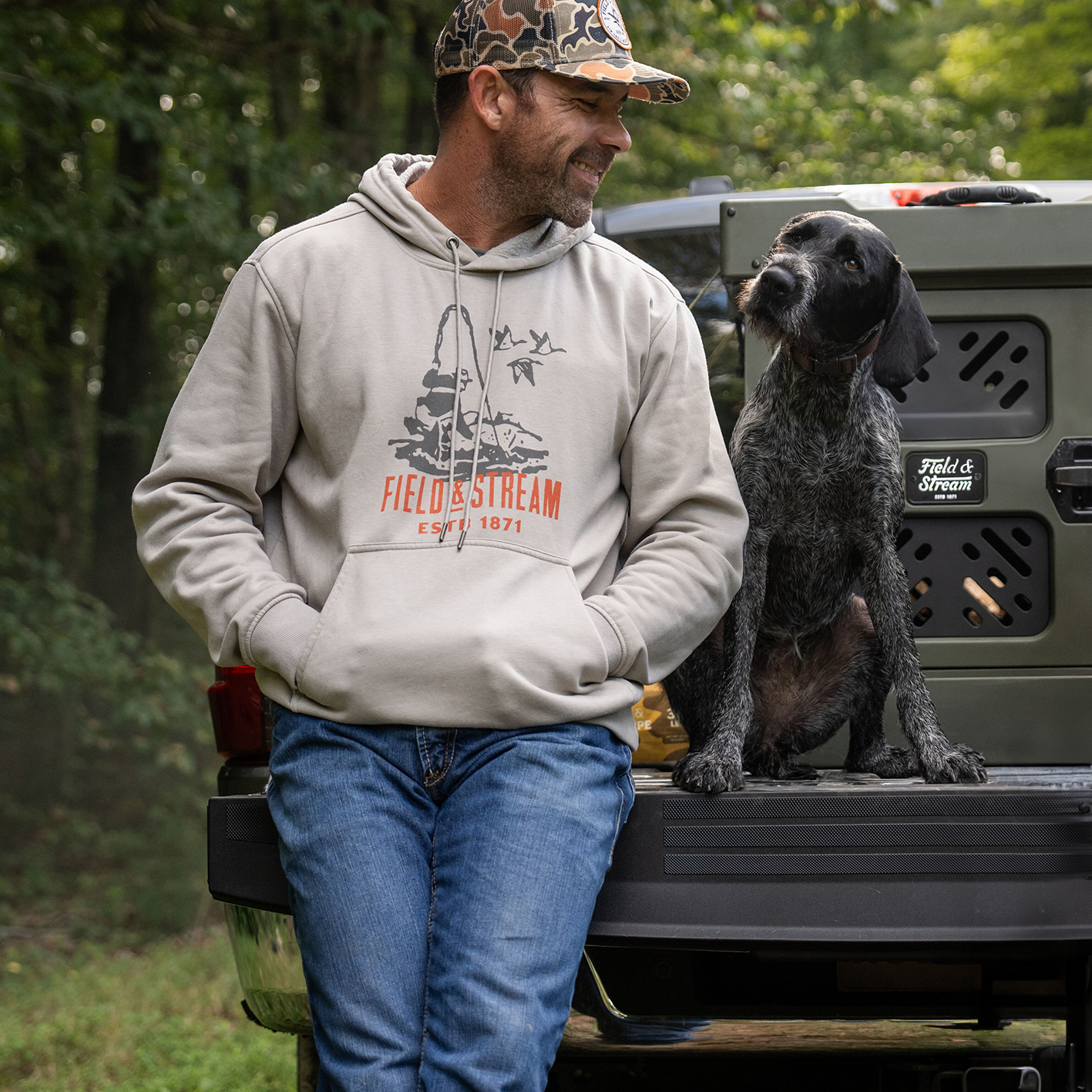 A man wearing a Field and Stream hoodie and camouflage baseball cap next to a dog on truck with a collapsible travel dog kennel in the background. See and shop items. 