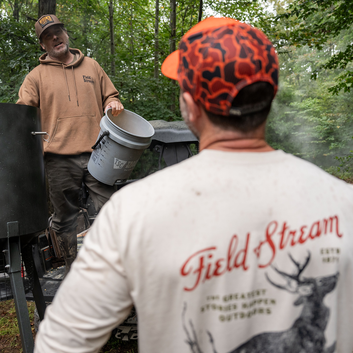 Two men talking outside, one wearing a Field and Stream hoodie and the other wearing a long-sleeve t-shirt with a Field and Stream logo and a deer printed on it. See and shop items.