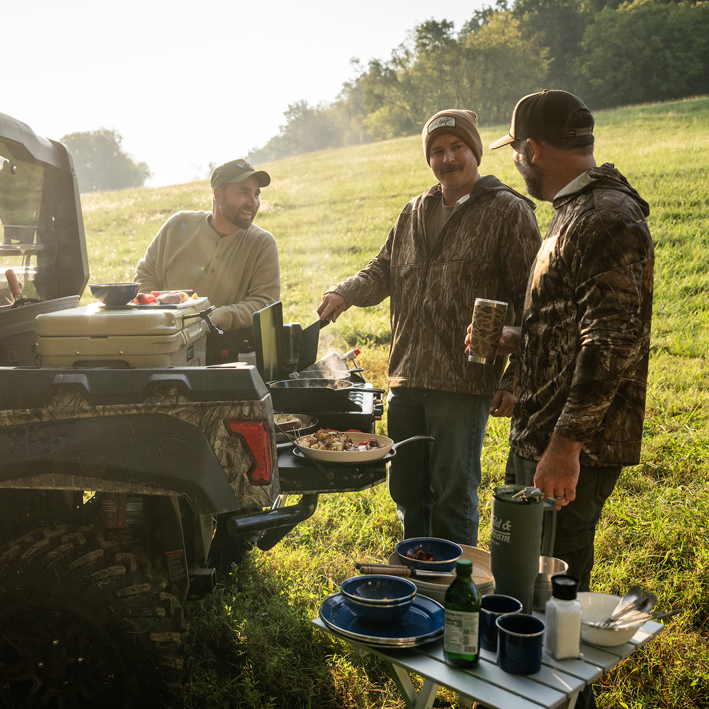 Three men wearing Field & Stream outdoor apparel are shown gathered around a grill.