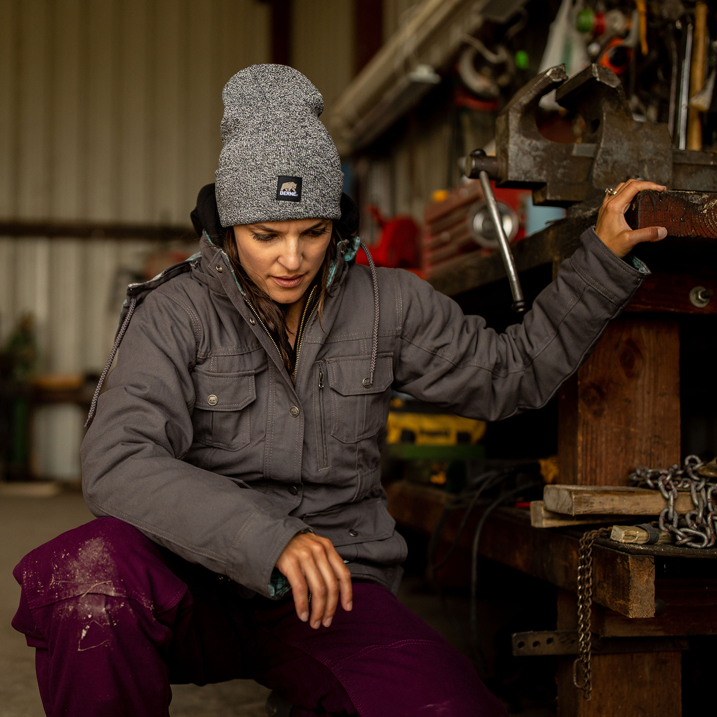 Woman in mechanic shop with grey heather hat and heavy work clothes. See and shop items. 