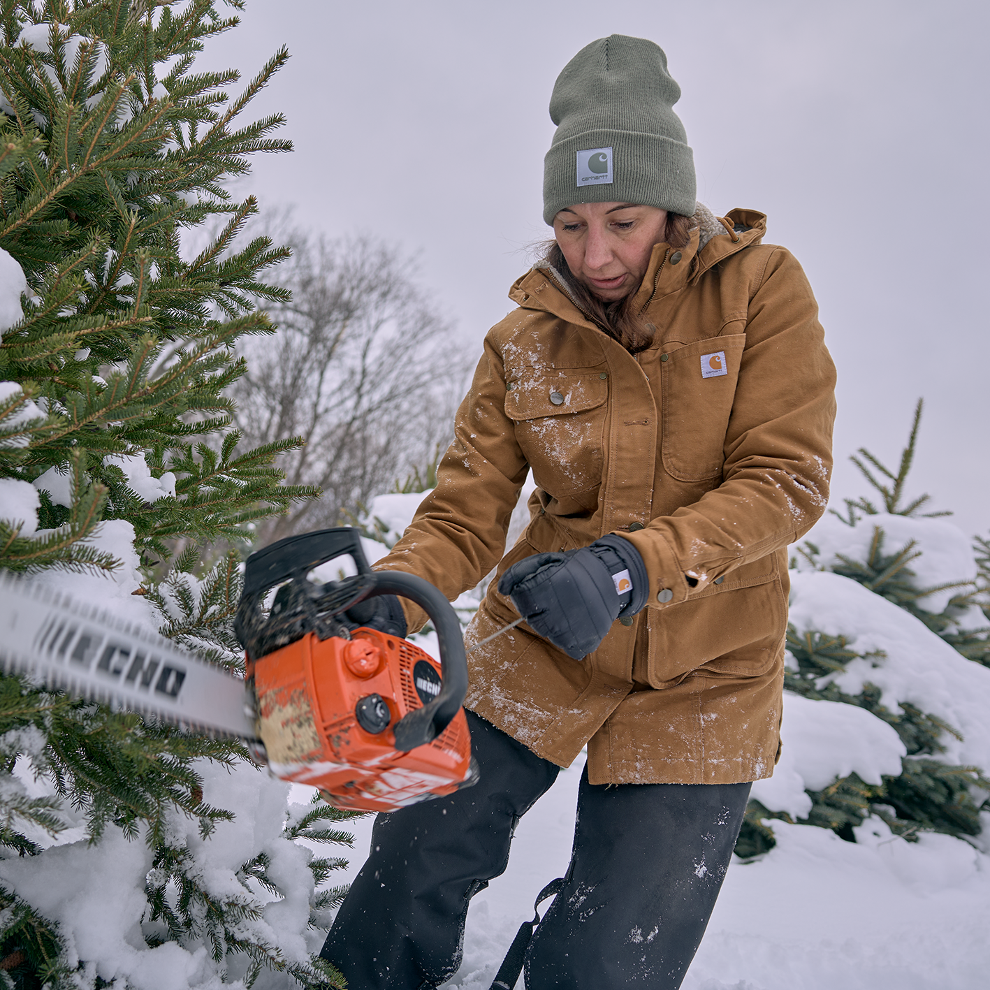 Woman in snow with chainsaw and warm clothes including hat, coat and gloves. See and shop items. 
