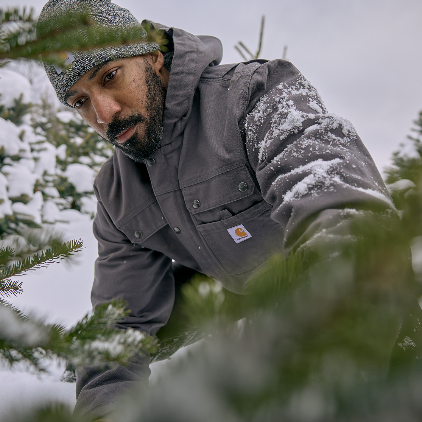 Man wearing a stocking cap and coat in snow behind fir tree. See and shop items. 
