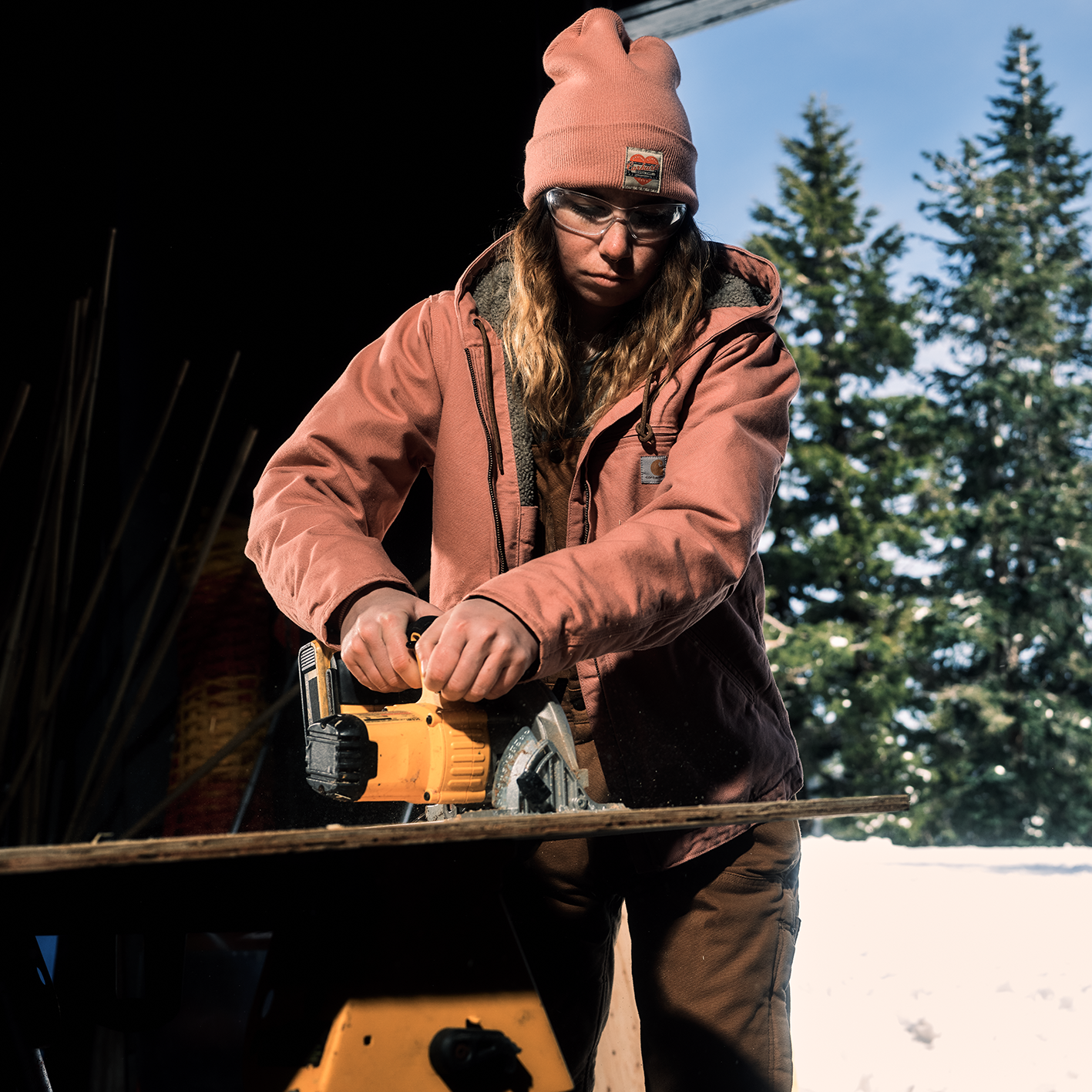 Woman cutting wood with power saw wearning a knit hat and heavy coat. See and shop items. 