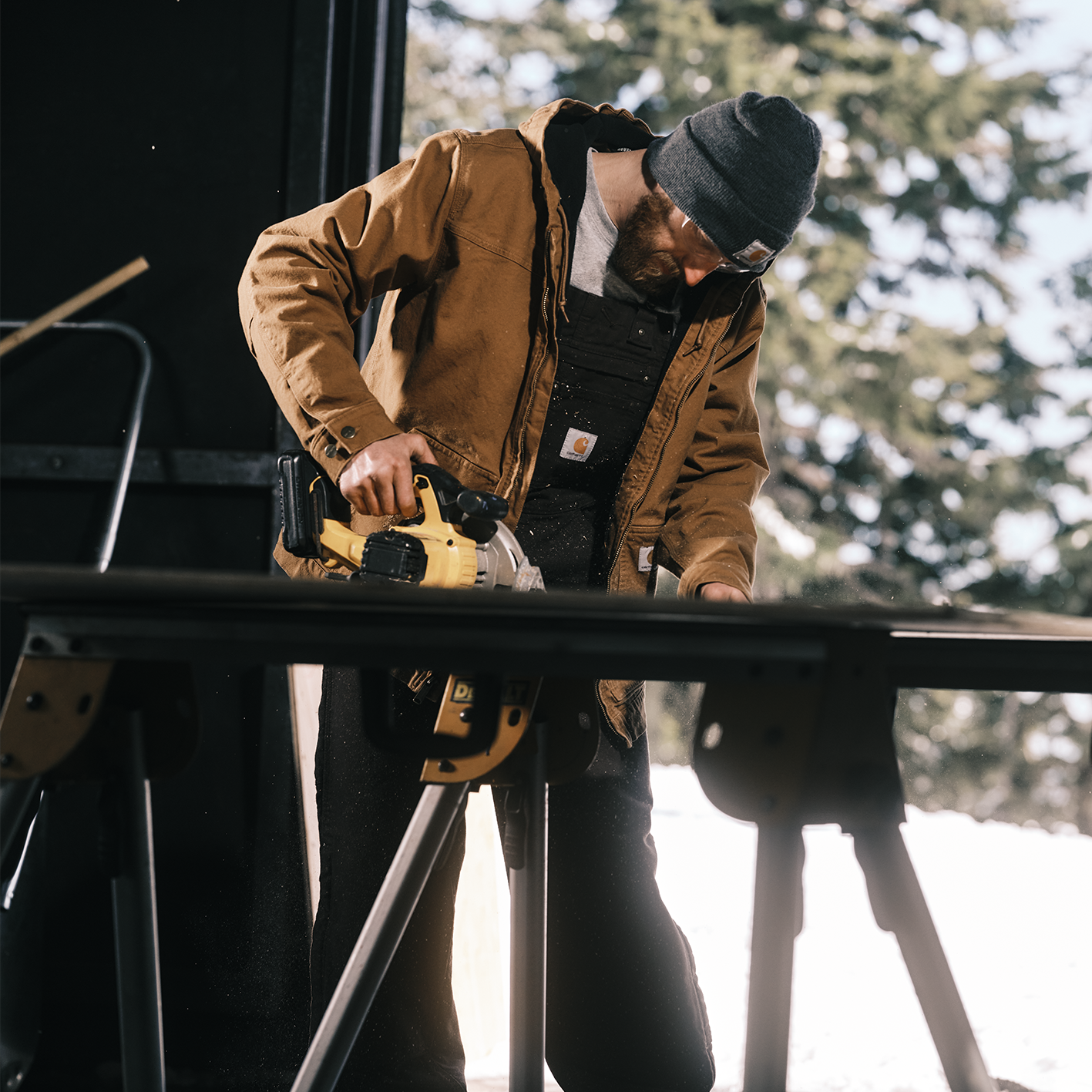 Man cutting wood with power saw wearing knit hat, heavy coat and coveralls. See and shop items. 