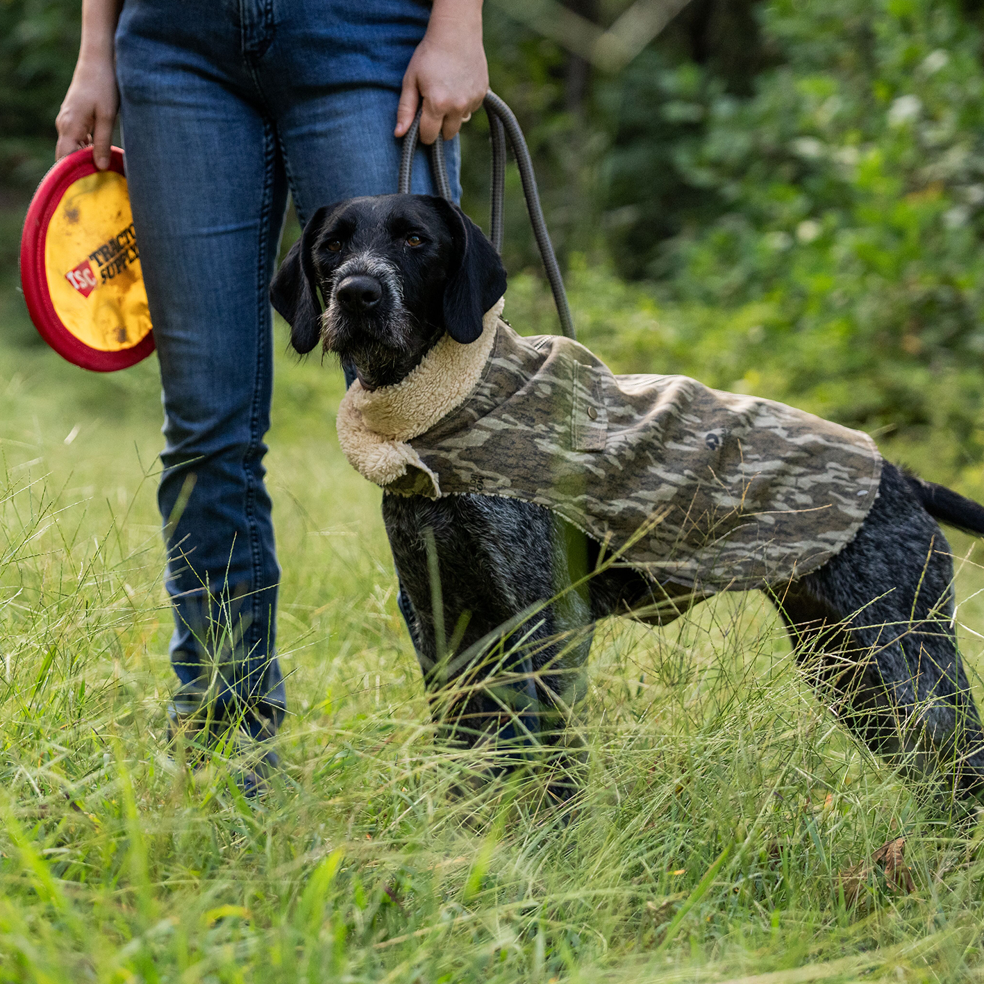 A man in a field with a large dog wearing a Field and Stream camouflage dog coat.