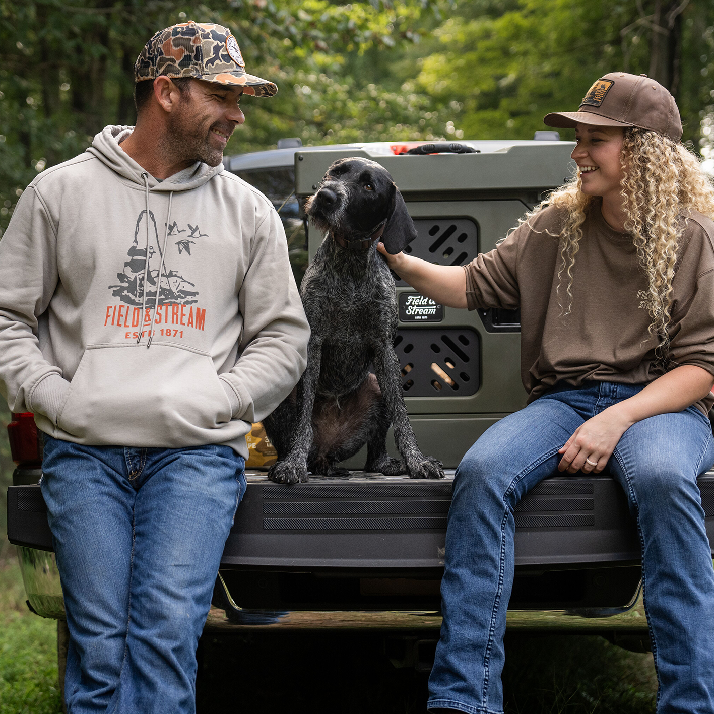A large dog with two people wearing Field and Stream clothing sitting on a truck bed with a dog next to a collapsible travel dog kennel.